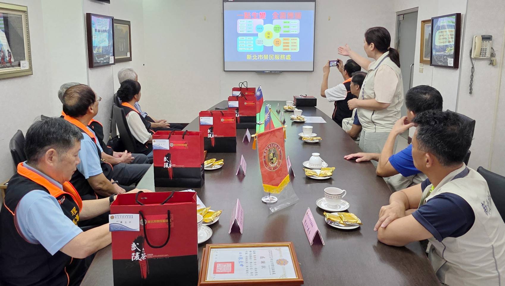 ▲新北市榮民服務處總幹事吳曉慧(站立者)向國防管理學院校友會理事長張永剛(左一)等一行人,介紹輔導會「安心御老平台」專案。/新北市榮服處提供。